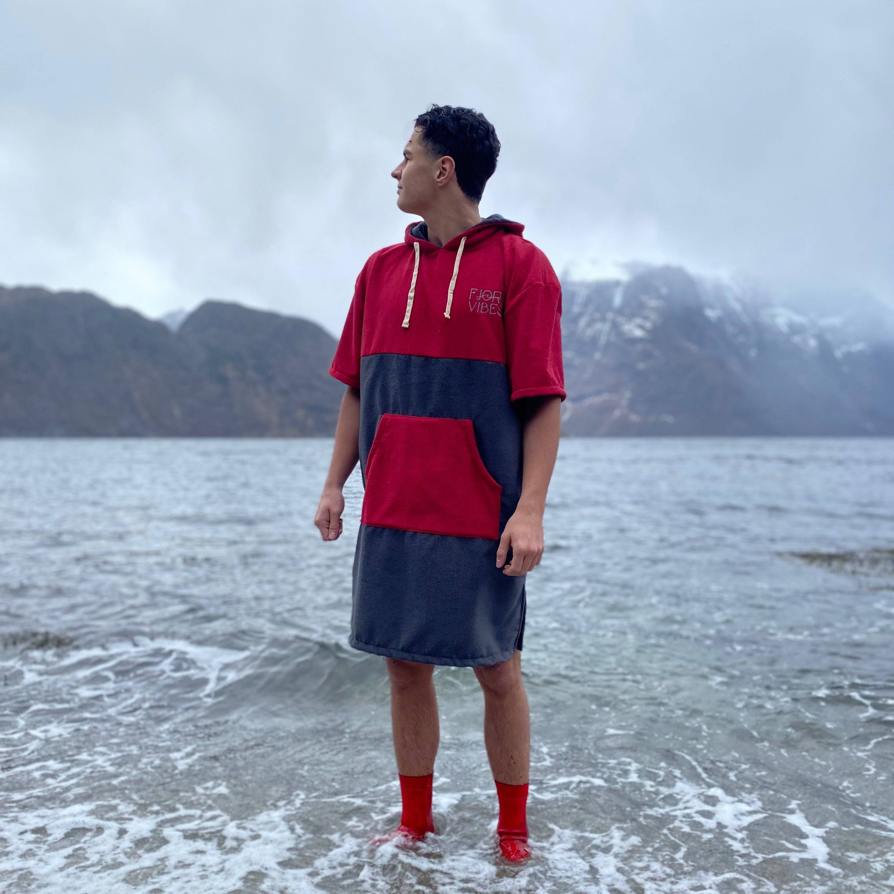 en mann som står i fjøra og ser mot fjellene. Han har på en rød og grå badeponcho for isbading