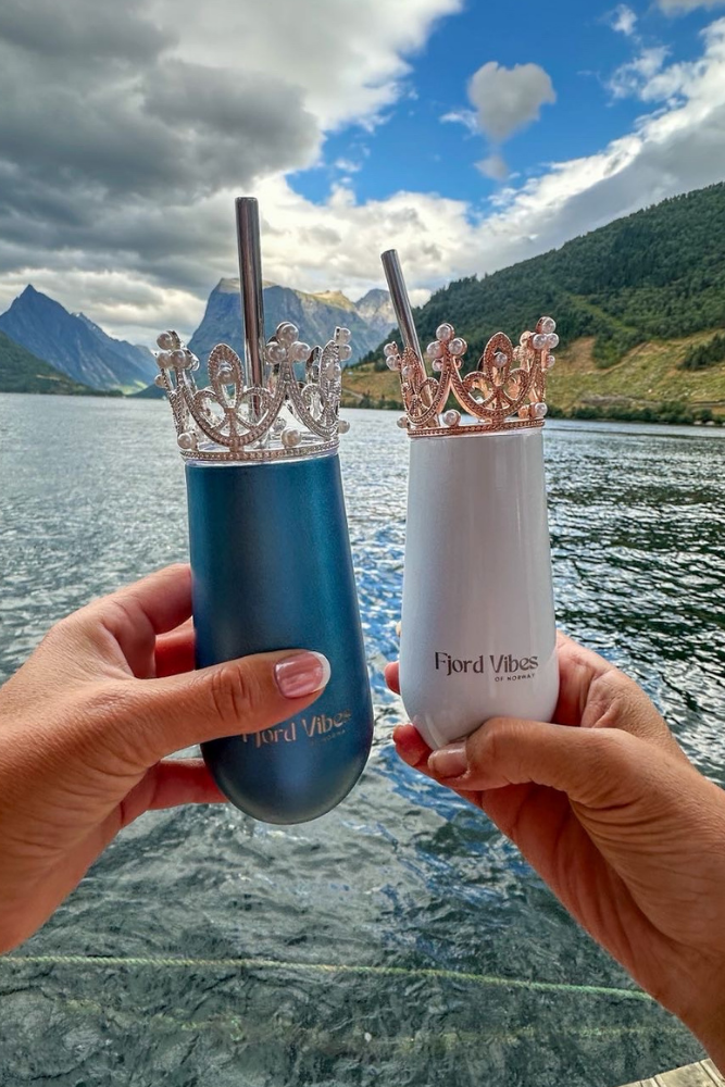 bilde av to hender som holder hver sin prosecco-kopp i hhv hvit og blå med her sin pyntekrone og sugerør. De skåler foran fjord og fjell