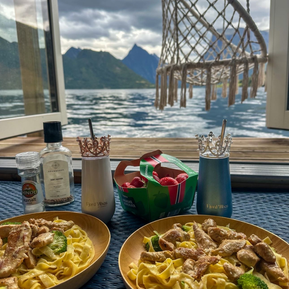 to middagstallerkener i vinduet mot en fjord, med prosecco kopper fra Fjord Vibes i hhv blå og hvit med hver sin pyntekrone og sugerør